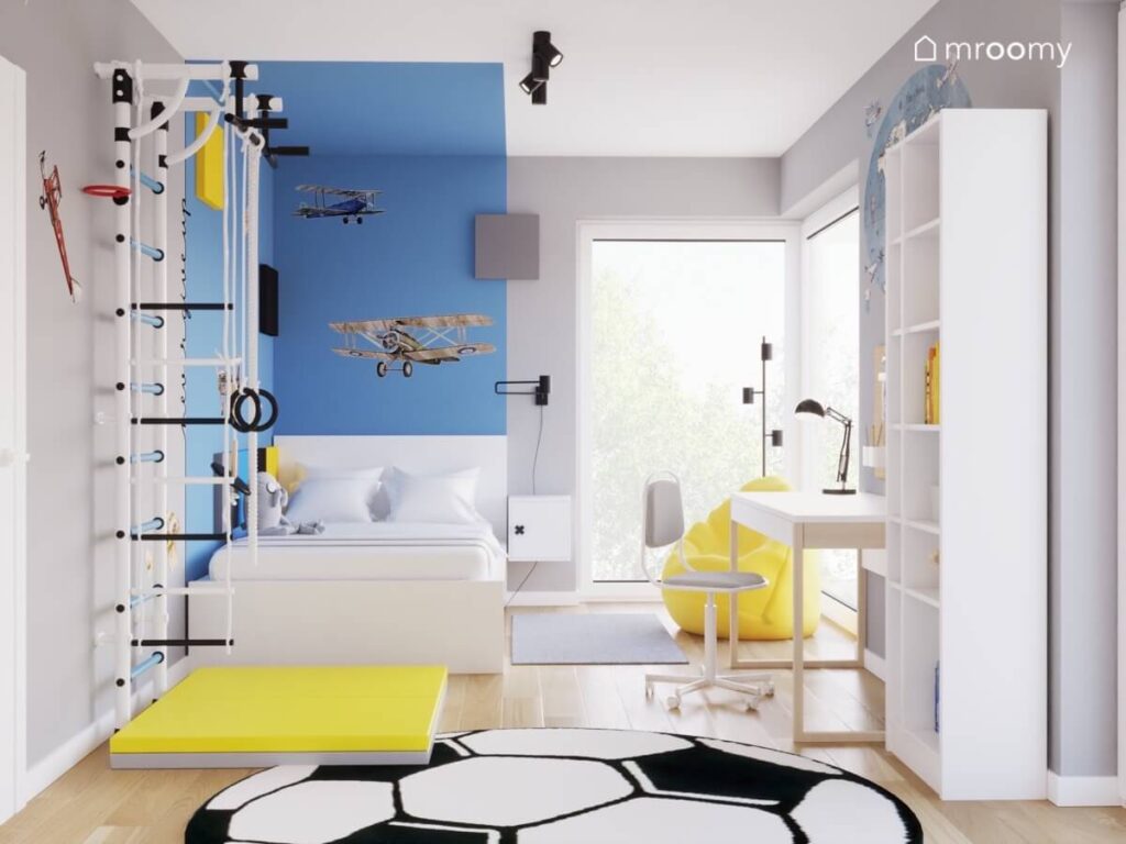 Szaro niebiesko biały pokój chłopca w wieku szkolnym a w nim białe łóżko regał drabinka gimnastyczna żółty materac oraz dywan w kształcie piłki nożnej