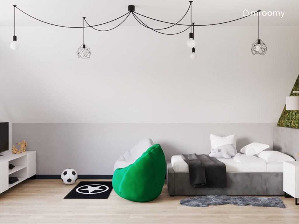 Biało szara ściana w pokoju nastoletniego chłopca pod nią szare tapicerowane łóżko oraz strefa rozrywki z telewizorem i dwoma pufami a na suficie lampy na rozłożystym zawieszeniu