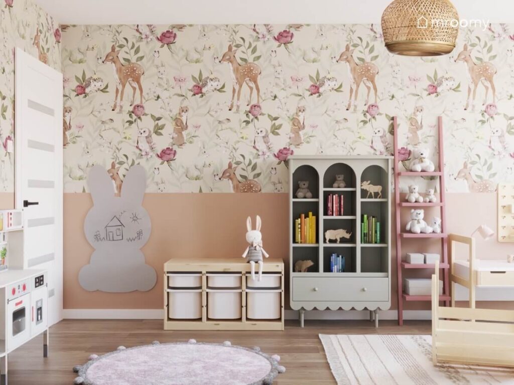 Przytulny pokój dwóch małych sióstr a w nim ściany pokryte tapetą w zwierzątka leśne i kwiaty tablica kredowa w kształcie królika drewniany regał z pojemnikami szary regał oraz różowy regał drabinka