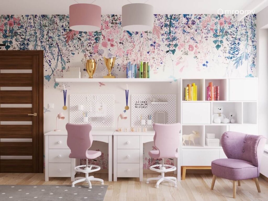 Strefa nauki w pokoju trzech dziewczynek a w niej biurka organizery półka różowe krzesła regał z szafkami a na ścianie tapeta w różowe i niebieskie kwiaty