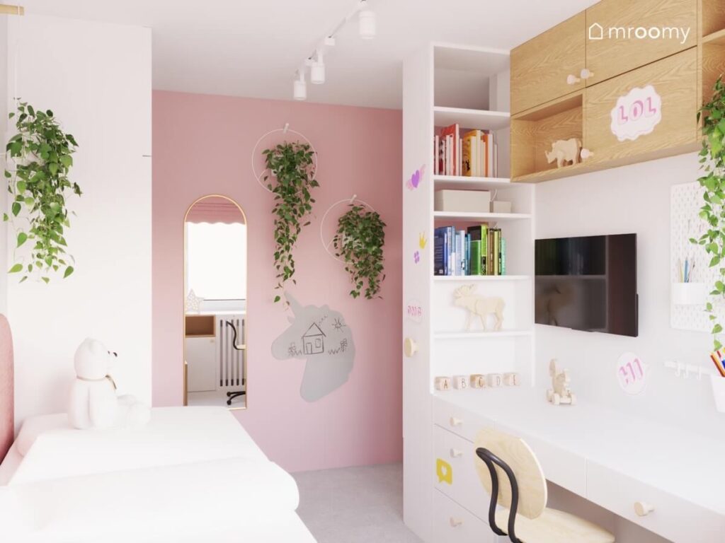 Biało różowy pokój dziewczynki w wieku szkolnym a w nim białe i drewniane meble liczne kwietniki tablica kredowa w kształcie jednorożca oraz duże lustro