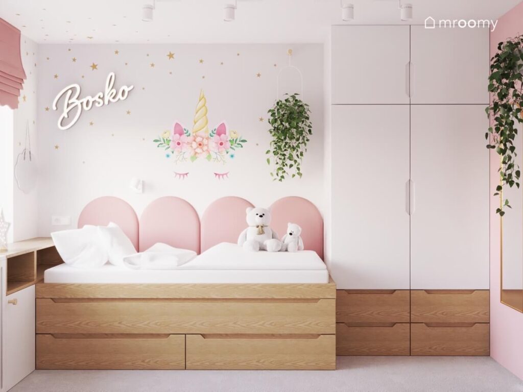 Drewniane łóżko w pokoju dziewczynki uzupełnione miękkimi panelami obok biało drewniana szafa a na ścianie ledon w kształcie napisu kwietnik i naklejka z jednorożcem