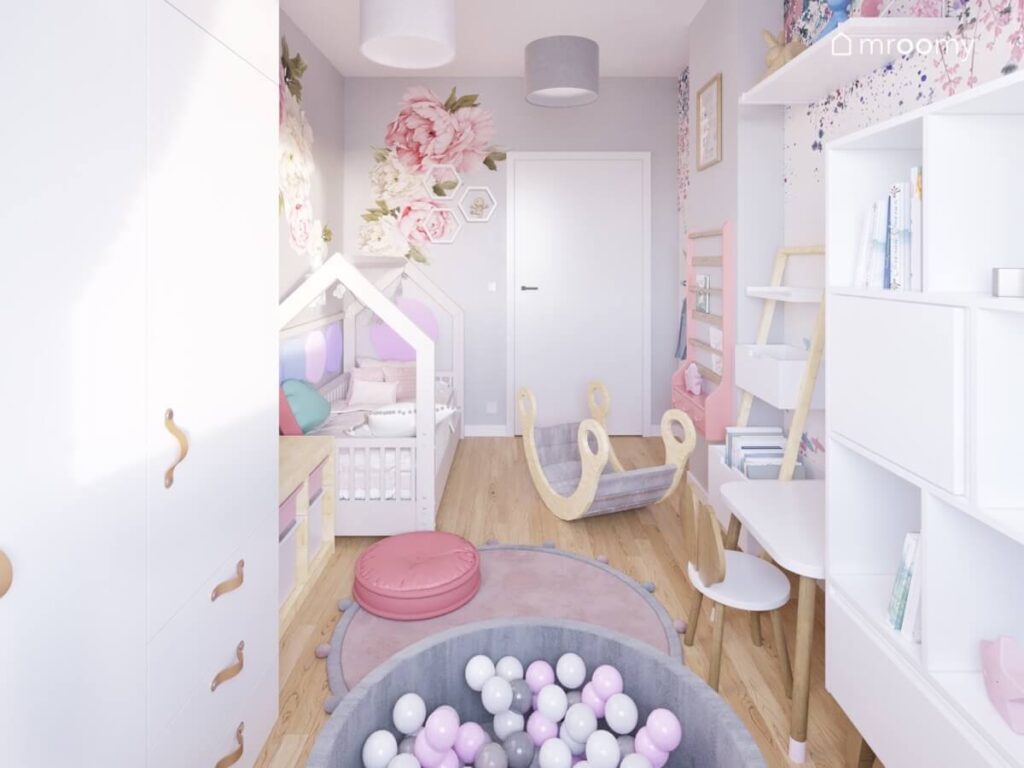 Biało szary pokój małej dziewczynki z łóżkiem domkiem bujakiem miękkim dywanem i pufami basenem z kulkami oraz naklejkami w kwiaty na ścianach