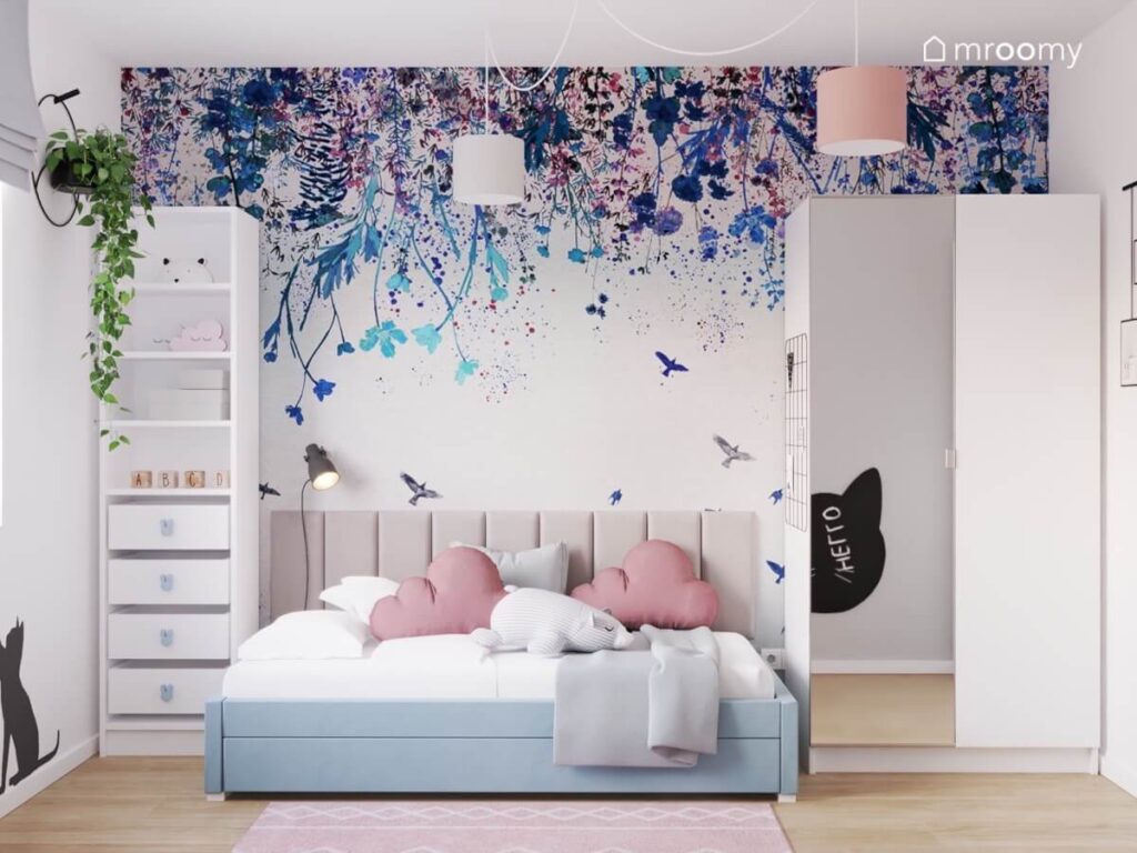 Strefa spania w pokoju dziewczynki a w niej niebieskie łóżko uzupełnione szarymi panelami i różowymi poduszkami obok wysoki regał i szafa z lustrzanym frontem a na ścianie tapeta w kwiaty