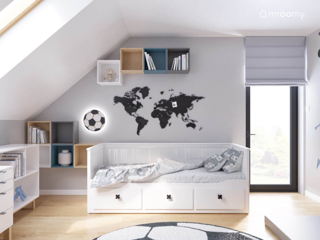 Szaro biały poddaszowy pokój dla chłopca w wieku szkolnym a w nim białe łóżko z szufladami a na ścianie liczne szafki mapa świata i lampka w kształcie piłki
