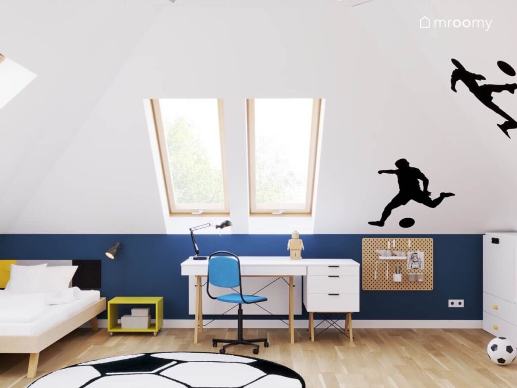 Poddaszowy pokój dla chłopca z białym skosem i granatową ścianką kolankową a także drewnianym łóżkiem białym biurkiem na drewnianych nogach i naklejkami w kształcie piłkarzy