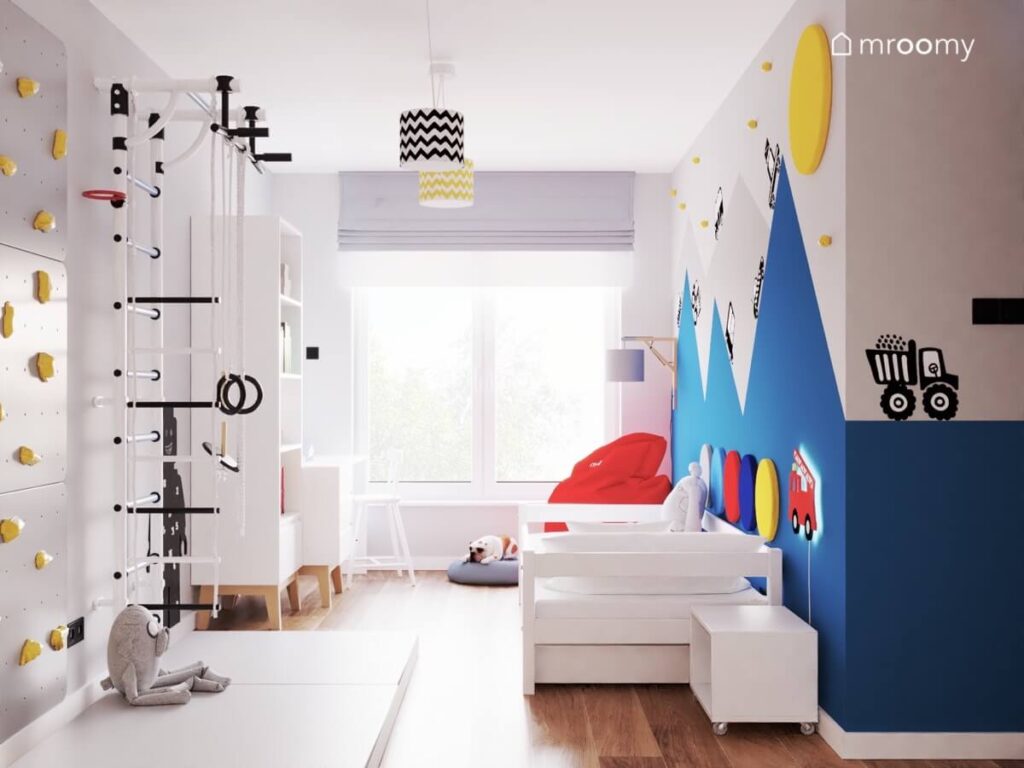 Jasny pokój dla małego chłopca a w nim białe meble drabinka gimnastyczna i ścianka wspinaczkowa a na ścianie malowanie w formie gór