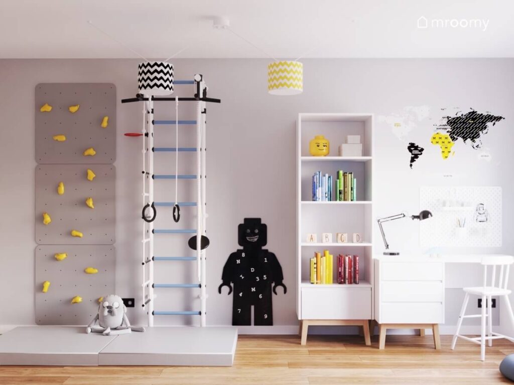 Szara ścianka wspinaczkowa z żółtymi panelami i drabinka gimnastyczna a obok tablica kredowa w kształcie ludzika Lego i wysoki regał w pokoju dla chłopca