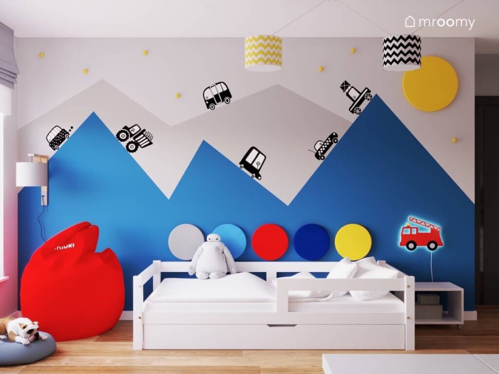 Białe łóżko a obok czerwona pufa a na ścianie szaro niebieskie malowanie w kształcie gór w pokoju trzylatka