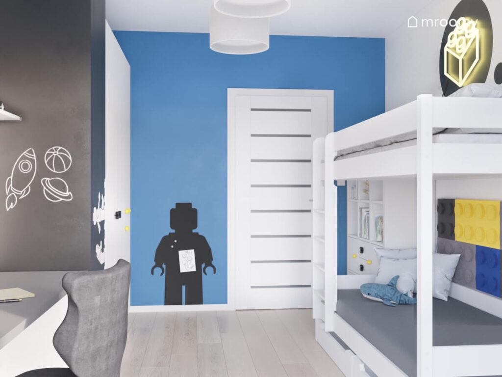 Powierzchnia kredowa niebieska ściana z tablicą w kształcie ludzika Lego oraz białe łóżko w pokoju kilkuletniego chłopca