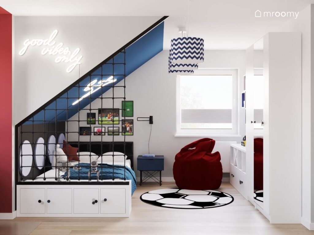 Poddaszowy pokój dla chłopca a w nim białe meble czarne łóżko bordowa pufa oraz dywan w kształcie piłki