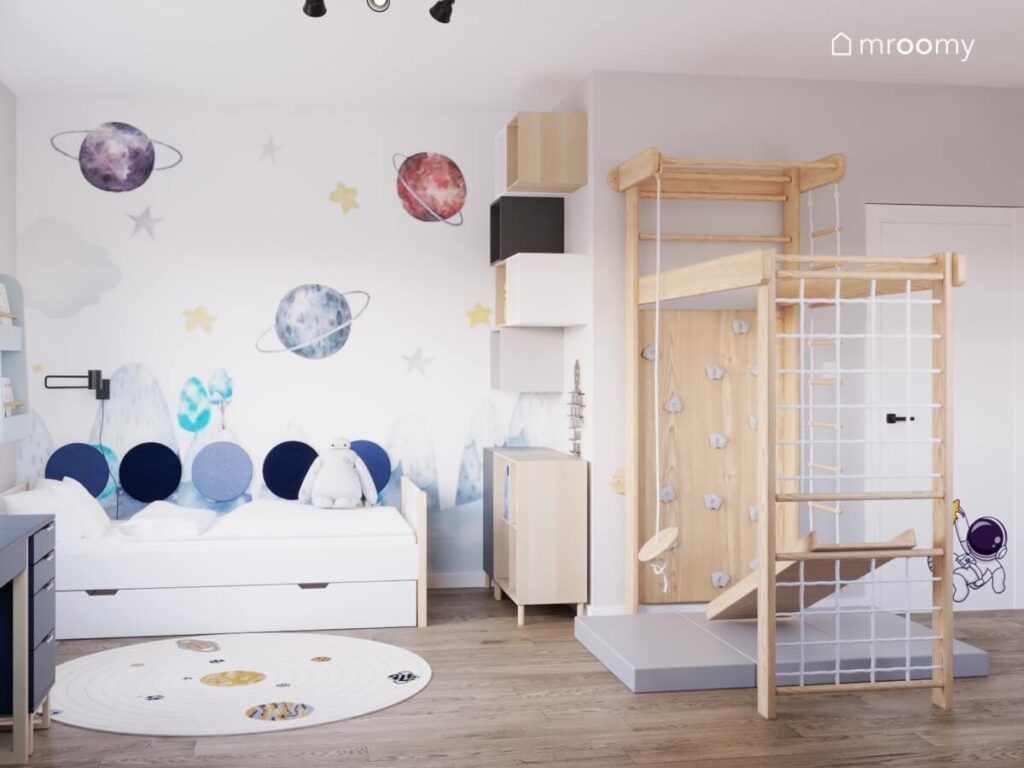 Białe łóżko uzupełnione panelami ściennymi w różnych odcieniach niebieskiego a obok drewniany plac zabaw ze ścianką wspinaczkową i siatką a na ścianie kosmiczna tapeta