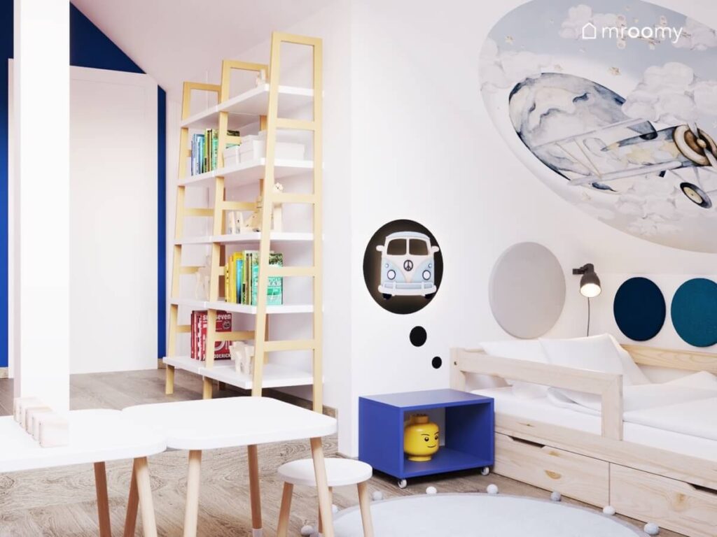 Jasny pokój dla dwóch małych chłopców na poddaszu a w nim biało drewniane regały na książki stoliki z krzesełkami oraz drewniane łóżko a na skosie naklejka z samolotem