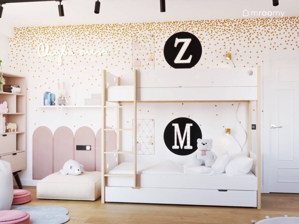 Biało drewniane łóżko piętrowe w pokoju dwóch sióstr w tym samym wieku a na ścianie inicjały dziewczynek tapeta w złote kropki oraz różowe panele
