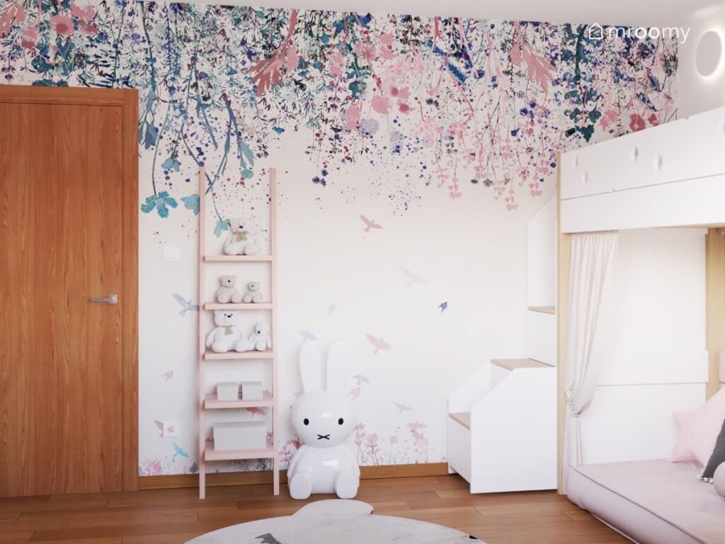 Ściana w pokoju siedmiolatki pokryta wzorzystą tapetą w kwiaty a na niej drewniany regał drabinka a obok niego lampka królik