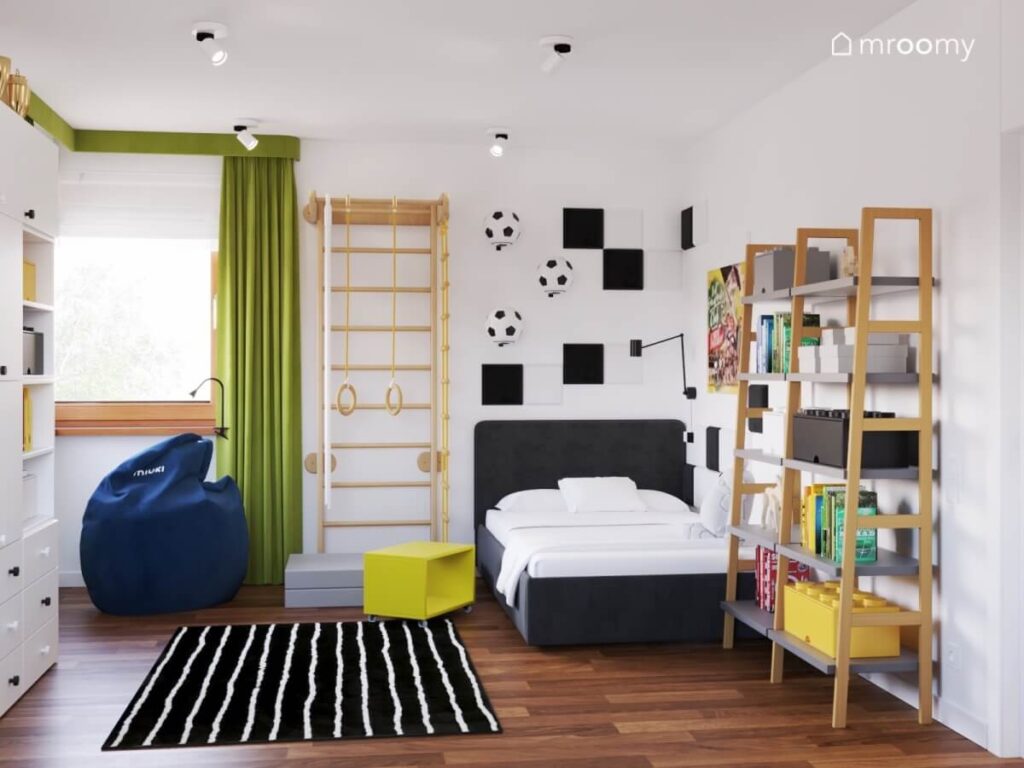 Ciemne tapicerowane łóżko uzupełnione białymi i czarnymi panelami ściennymi obok drewniana drabinka gimnastyczna a na podłodze dywan w paski w pokoju dla chłopca