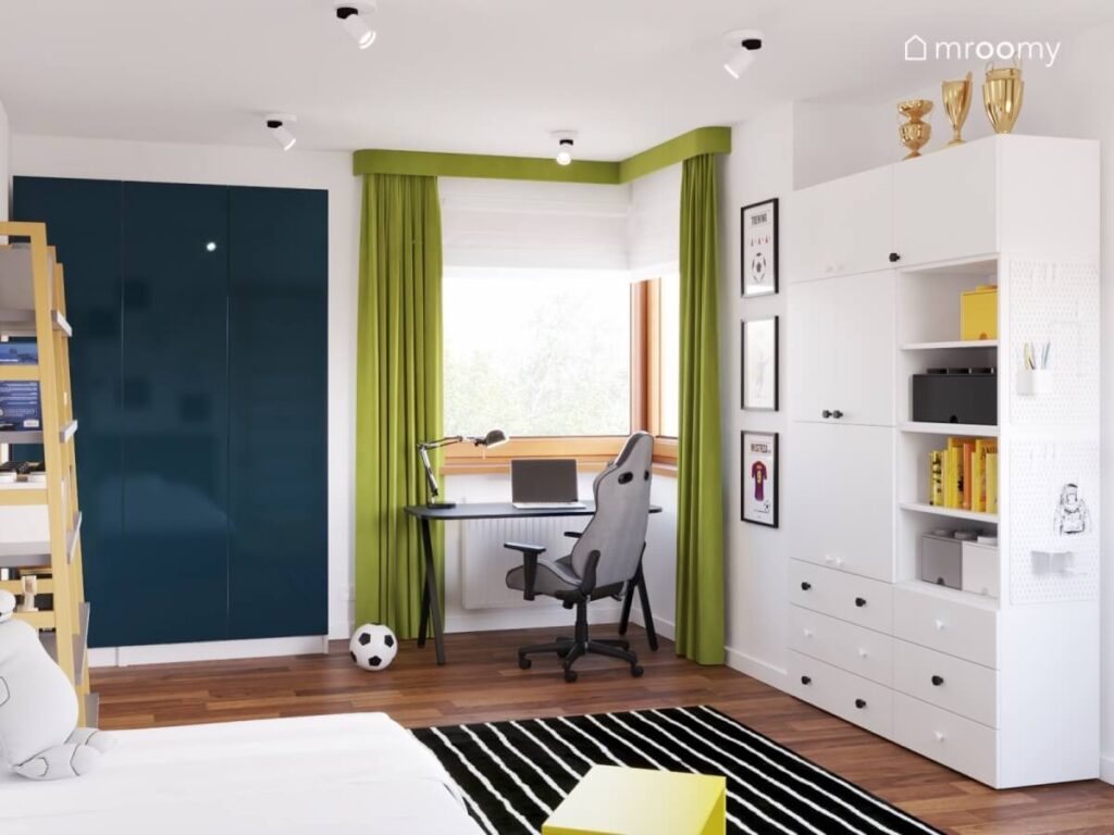 Biały pokój dla chłopca z granatową i białą szafą biurkiem pod oknem z zielonymi zasłonami oraz dywanem w paski