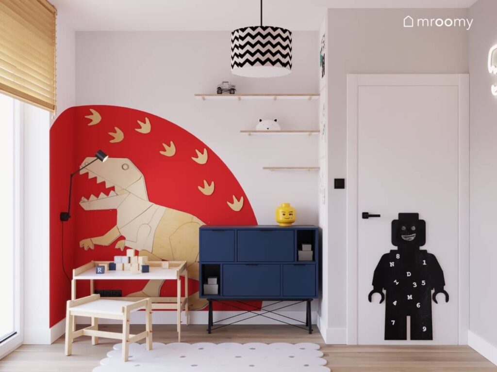 Dinozaur ze sklejki na czerwonym tle oraz granatowa komoda drewniany stolik i tablica kredowa ludzik Lego w pokoju dla chłopca