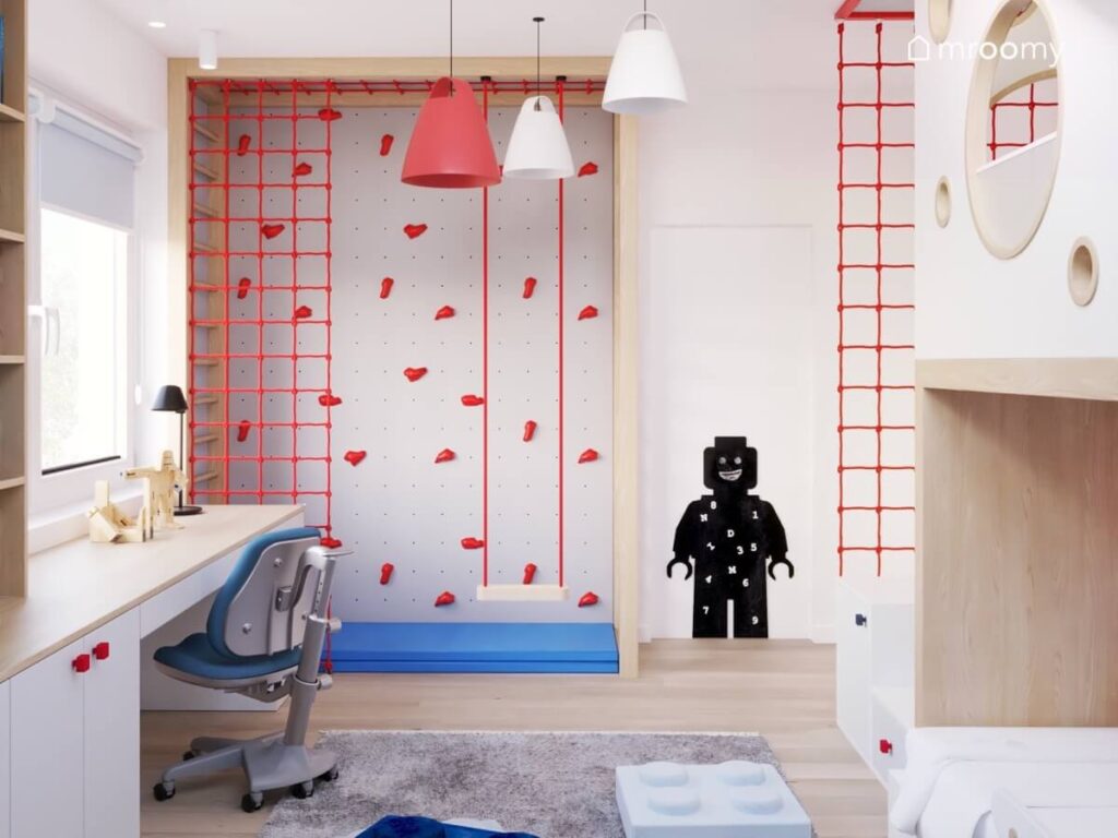 Strefa ruchu w pokoju kilkulatka a w niej szara ścianka wspinaczkowa z czerwonymi uchwytami siatką i niebieskim materacem oraz huśtawka wisząca a obok tablica kredowa w kształcie ludzika Lego na drzwiach