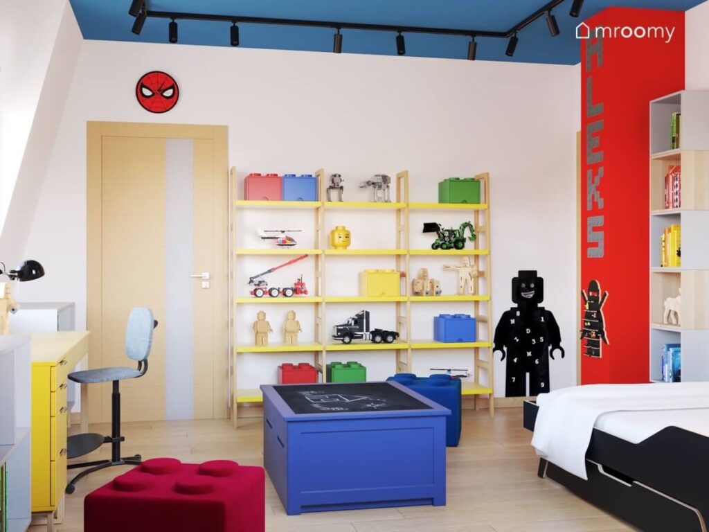 Biało niebiesko czerwony pokój dla chłopca a w nim regały w formie drabinki tablica kredowa w kształcie ludzika Lego niebieski stolik do zabawy oraz pufy klocki