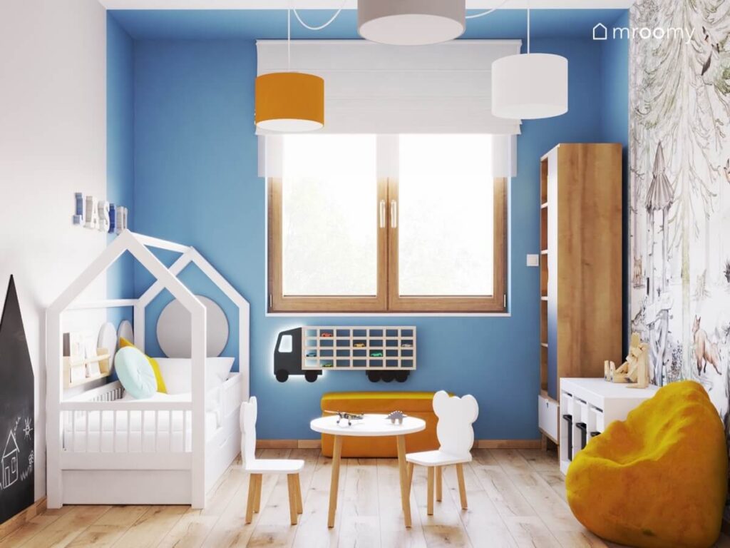 Niebiesko biały pokój chłopca a w nim białe łóżko domek stolik z krzesełkami w kształcie misiów półka na samochody oraz pomarańczowe dodatki