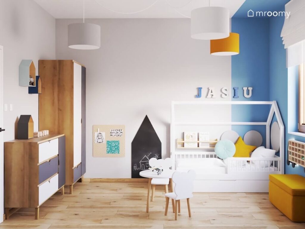 Szaro niebieski pokój małego chłopca a w nim drewniano biało granatowe meble tablica kredowa w kształcie domku oraz białe łóżko domek