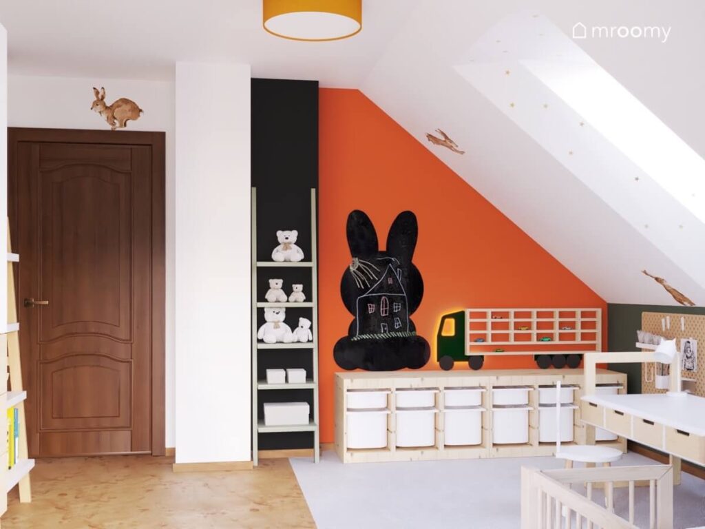 Pomarańczowa ściana w poddaszowym pokoju chłopca a na niej drewniany regał z pojemnikami półka na samochody w kształcie ciężarówki i tablica kredowa w kształcie królika