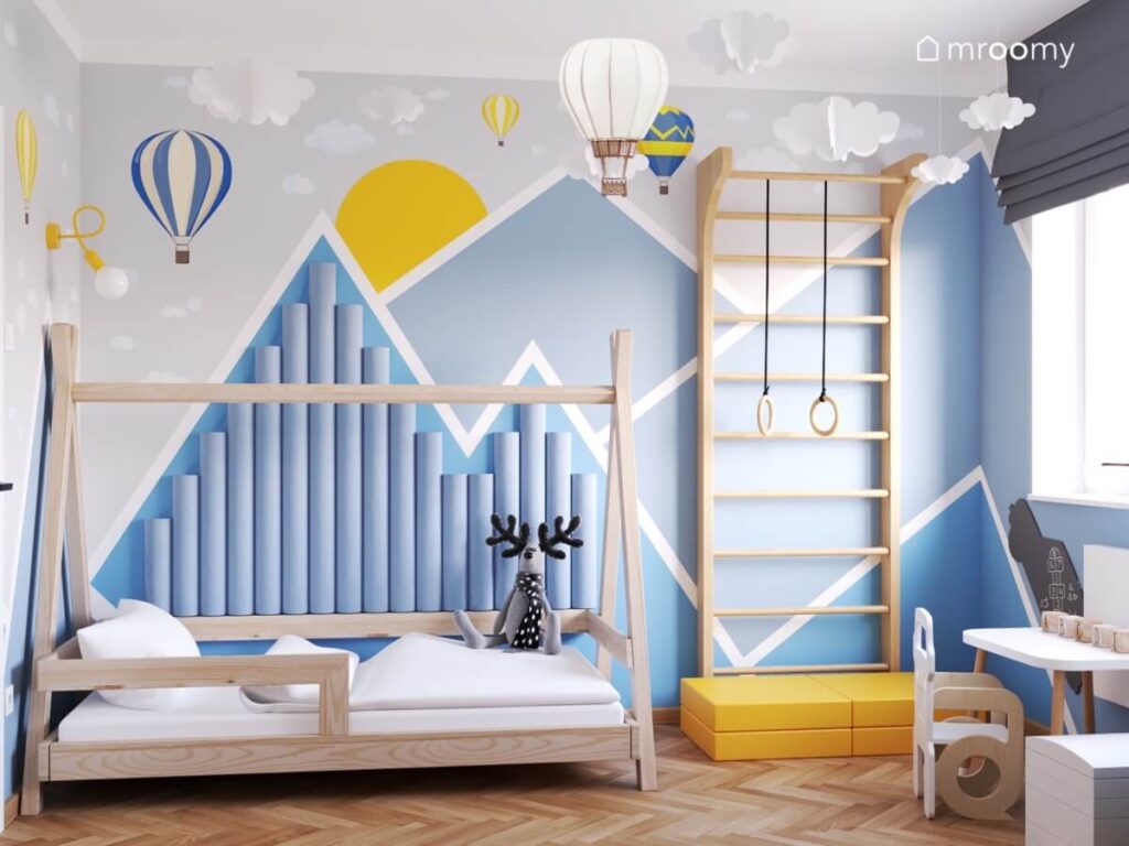 Niebieska geometryczna kompozycja na ścianie w pokoju chłopca a na niej drabinka gimnastyczna i łóżko tipi