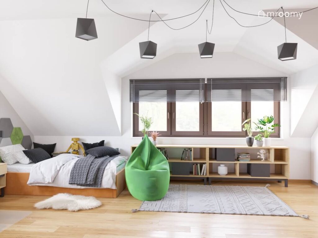 Jasny poddaszowy pokój dla chłopca a w nim pomarańczowe łóżko zielona pufa dwa dywany niskie regały oraz oryginalne geometryczne lampy sufitowe