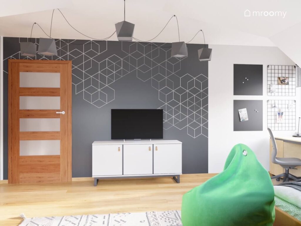 Ściana w pokoju chłopca pokryta geometrycznymi wzorami a na niej telewizor i szafka obok organizery ścienne a na suficie lampa na zawieszeniu typu pajączek