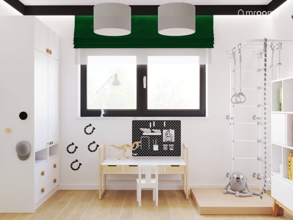 Biała szafa niski stolik z organizerem drabinka gimnastyczna z materacem oraz zielona roleta a na ścianie naklejki w kształcie podków w pokoju chłopca