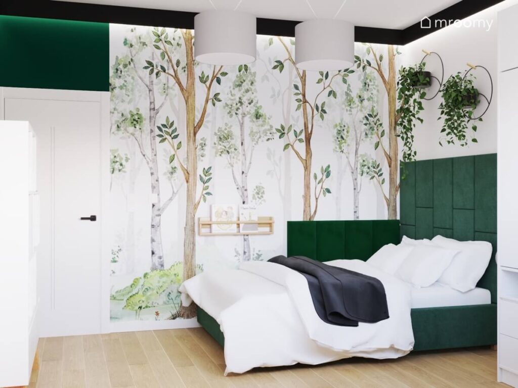 Strefa spania w pokoju kilkulatka a w niej ciemnozielone tapicerowane łóżko półka na książki dwa kwietniki oraz tapeta w drzewa
