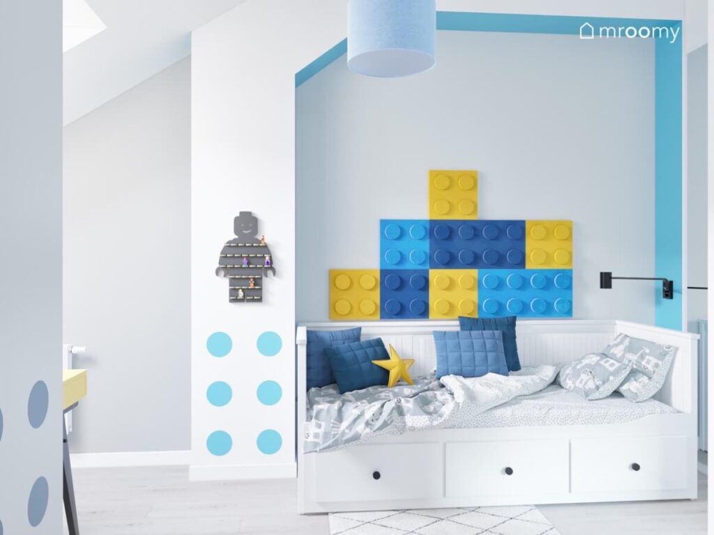 Strefa spania w pokoju dla chłopca a w niej białe łóżko z szufladami ściana uzupełniona kolorowymi panelami w kształcie klocków oraz półeczka w kształcie ludzika Lego
