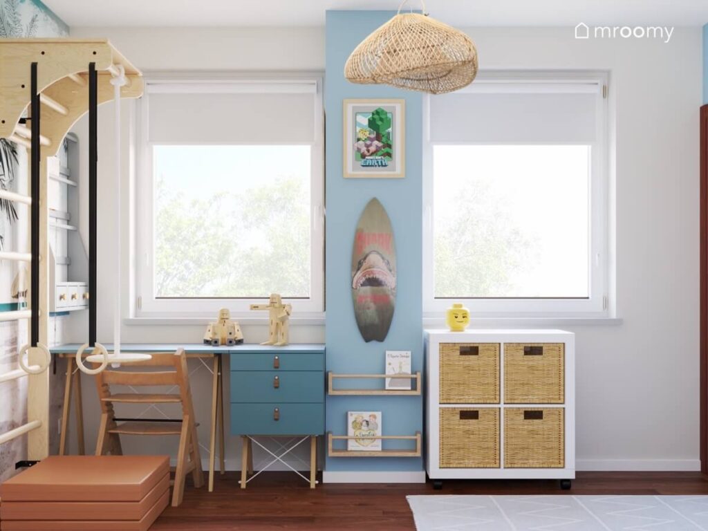 Niebieskie biurko z kontenerkiem i drewnianym krzesłem kwadratowy regał z pojemnikami a na środku filar z deską surfingową półeczkami i obrazkiem w pokoju chłopca