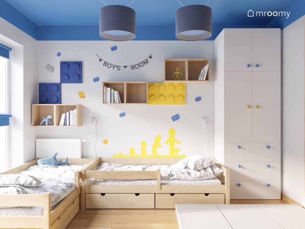 Jasny pokój dla kilkuletnich chłopców z niebieskim sufitem drewnianymi łóżkami i szafkami ściennymi oraz licznymi motywami Lego