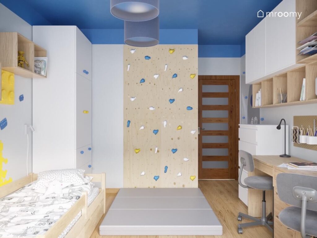Ścianka wspinaczkowa z kolorowymi uchwytami oraz szarymi materacami a także białe i drewniane meble w pokoju dwóch chłopców