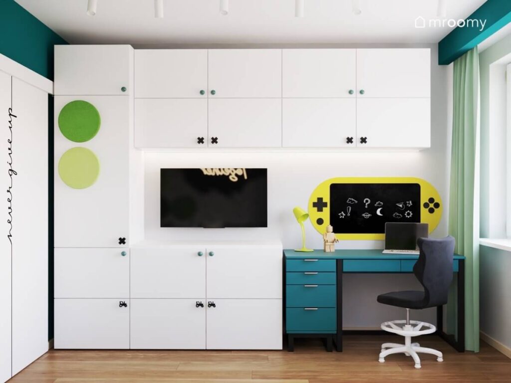Białe meble modułowe uzupełnione gałkami w różnych kształtach i zielonymi panelami a także niebieskie biurko i gamingowa tablica kredowa w pokoju dla chłopców