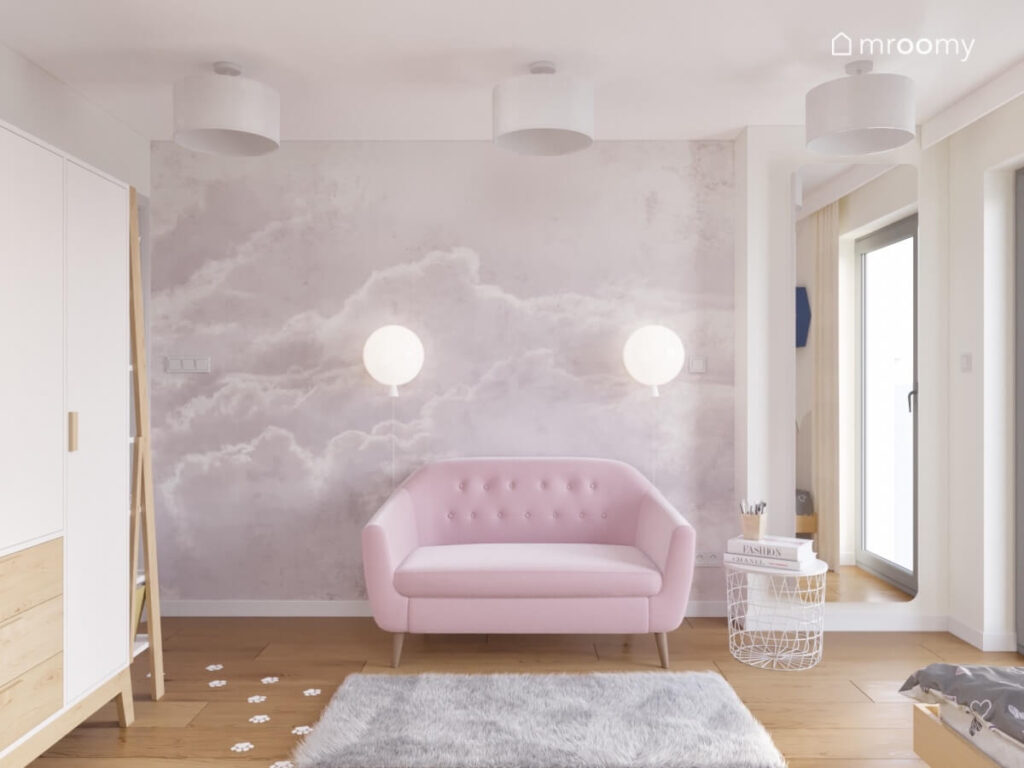 Ściana pokryta tapetą w chmury a pod nią różowa sofa oświetlana przez kinkiety balony w białym pokoju dziewczynki