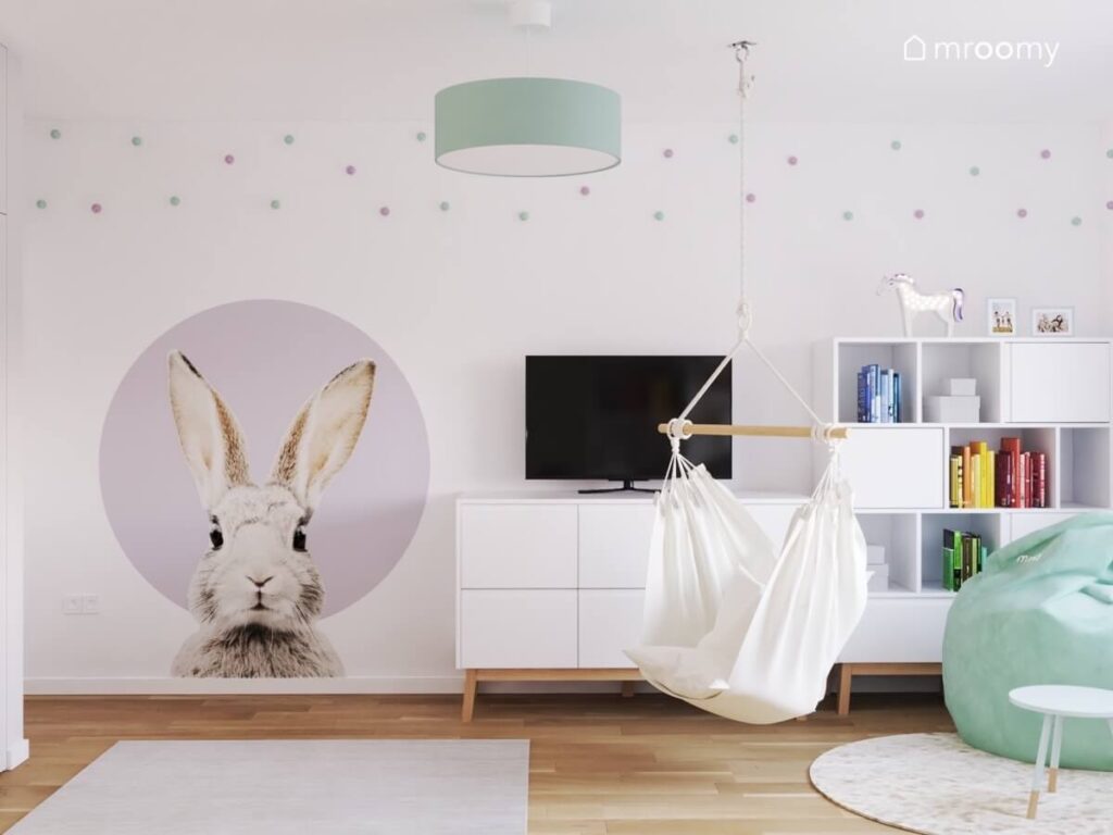 Biała ściana w pokoju dziewczynki a na niej naklejka z królikiem szafka rtv z telewizorem biały regał oraz ozdobne fioletowe i miętowe gałki