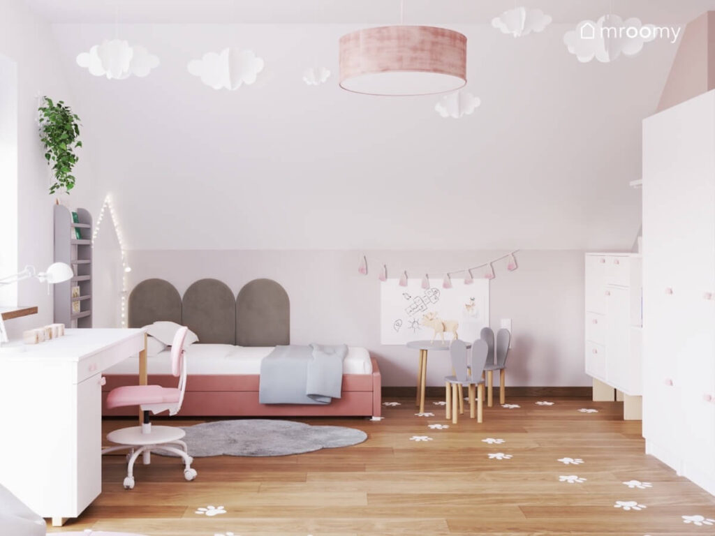 Różowe łóżko z ozdobnym oświetleniem w formie domku i szarymi panelami obok białe biurko i stolik z krzesełkami w kształcie króliczych uszu w poddaszowym pokoju dziewczynki