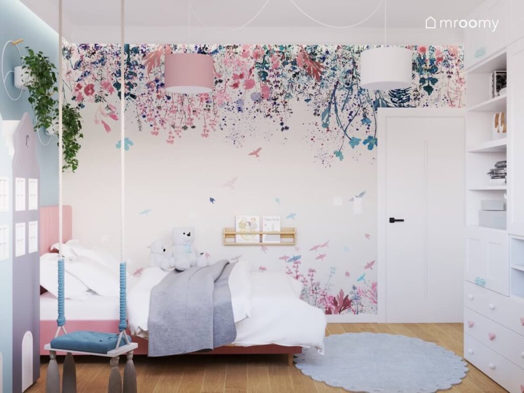 Ściana w pokoju dla dziewczynki pokryta tapetą w różowe i niebieskie kwiaty a pod nią różowe łóżko