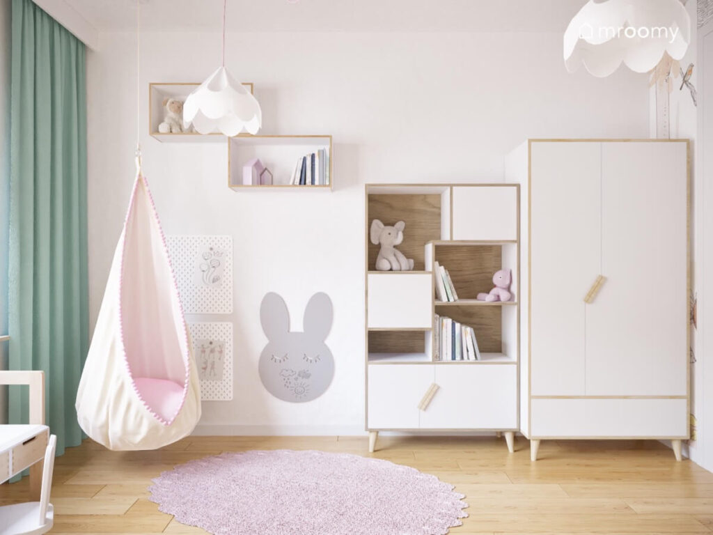 Biało drewniane meble szara tablica kredowa w kształcie królika wiszący fotel kokon oraz dywan w pokoju rocznej dziewczynki