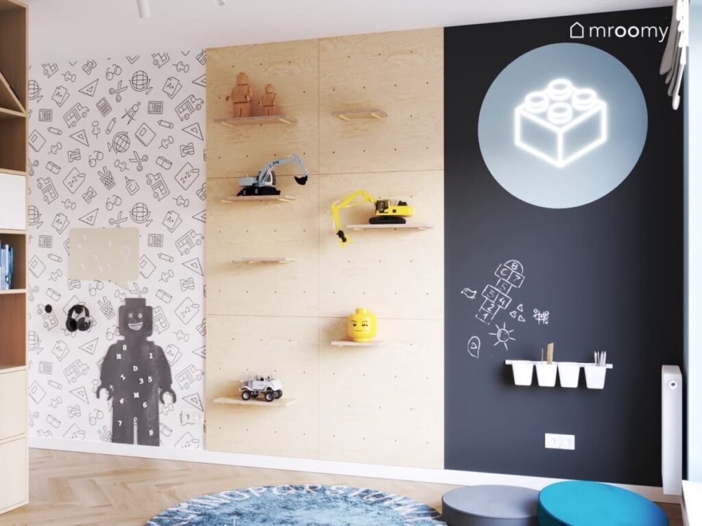 Ściana w pokoju dla rodzeństwa z tapetą w naukowy wzór półkami na zabawki powierzchnią kredową oraz tablicą magnetyczną w kształcie ludzika Lego i ledonem klocek