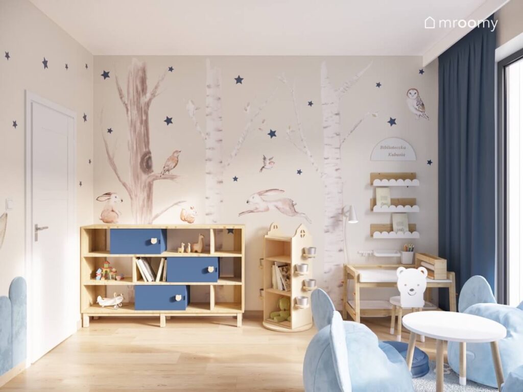Beżowa ściana w pokoju małego chłopca pokryta leśnymi naklejkami z drzewami i zwierzętami a także granatowymi gwiazdkami a na niej drewniane meble