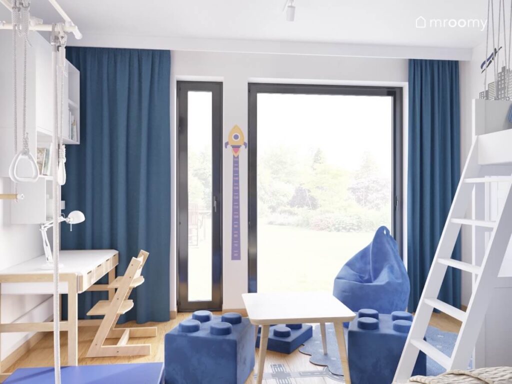 Jasna ściana w pokoju chłopca a na niej okna z ciemnoniebieskimi zasłonami oraz kosmiczna miarka wzrostu a na podłodze niebieskie pufy i stolik