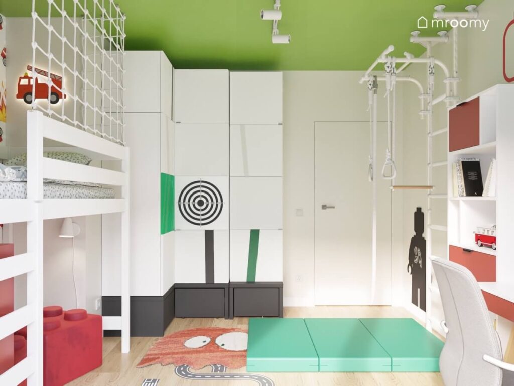 Biała szafa z czarnym podestem oraz ozdobnymi frontami a na ścianie obok drabinka gimnastyczna i materac w szarym pokoju dla chłopca z zielonym sufitem