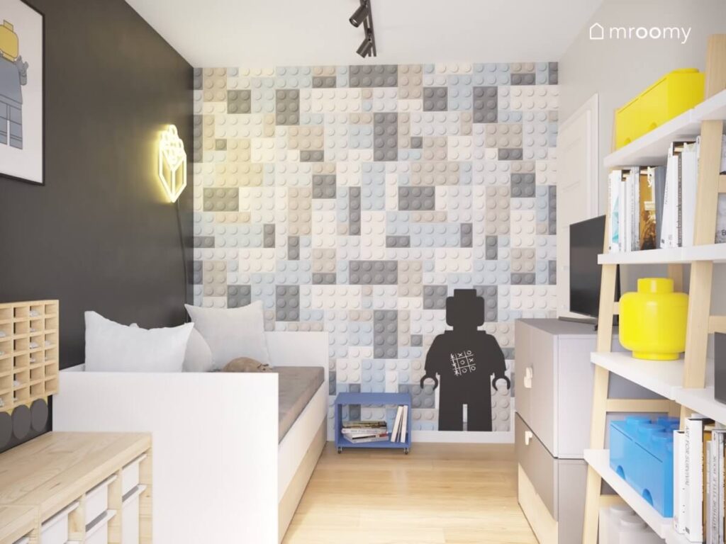 Ściana pokryta tapetą w klocki Lego a na niej tablica kredowa w kształcie ludzika w czarno szarym pokoju dla chłopca z biało drewnianymi meblami