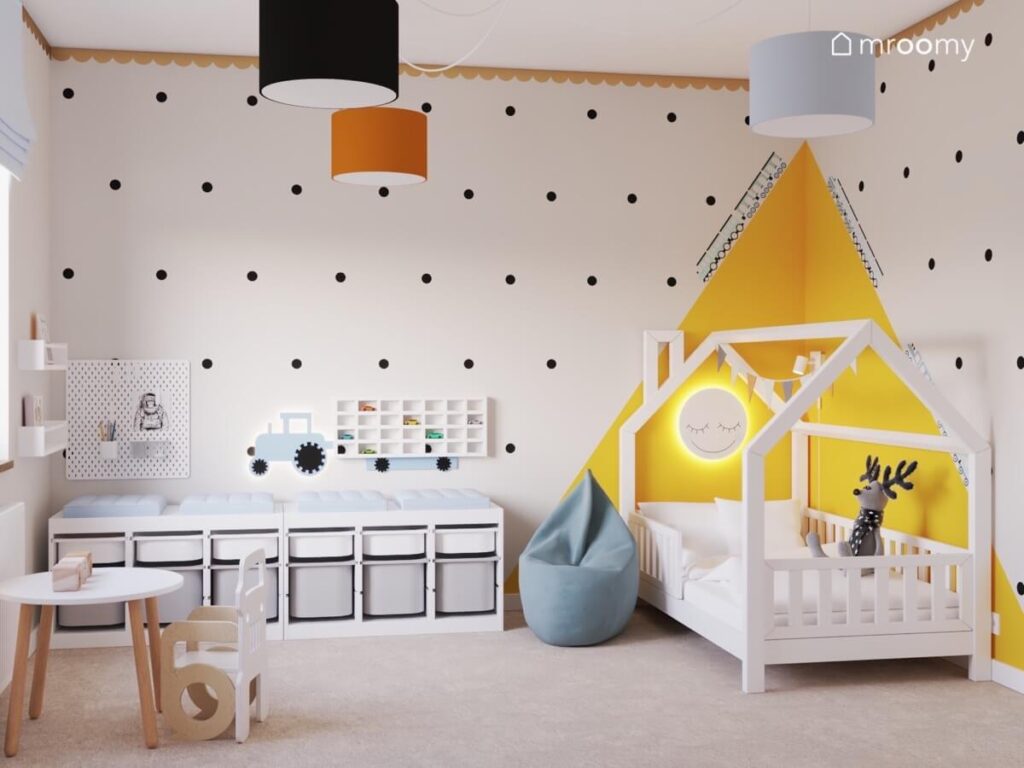 Jasny przytulny pokój dla chłopca a w nim regał z pojemnikami i łóżko domek na ścianie kropki a na suficie pomarańczowa czarna i biała lampa