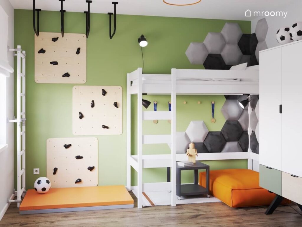 Zielona ściana w pokoju chłopca a na niej panele wspinaczkowe oraz miękkie panele w kształcie sześciokątów a oprócz tego antresola z miejscem do spania i pomarańczowa pufa i materac