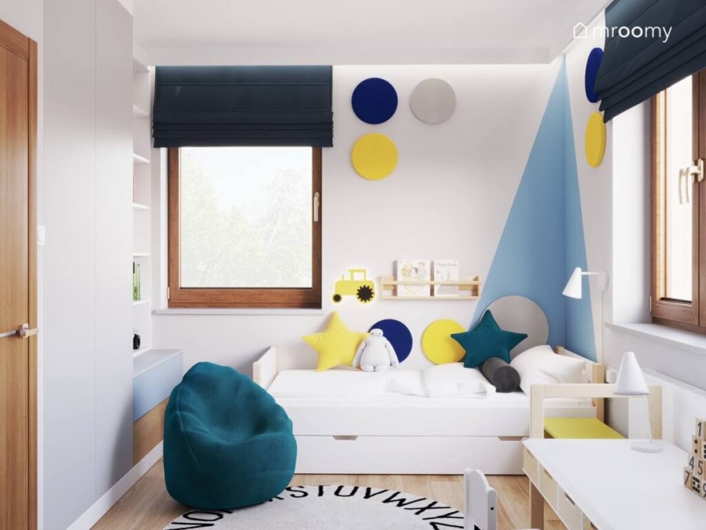 Strefa spania w pokoju chłopca w niej proste łóżko z kolorowymi poduszkami powyżej panele ścienna a na podłodze pufa oraz dywan z alfabetem